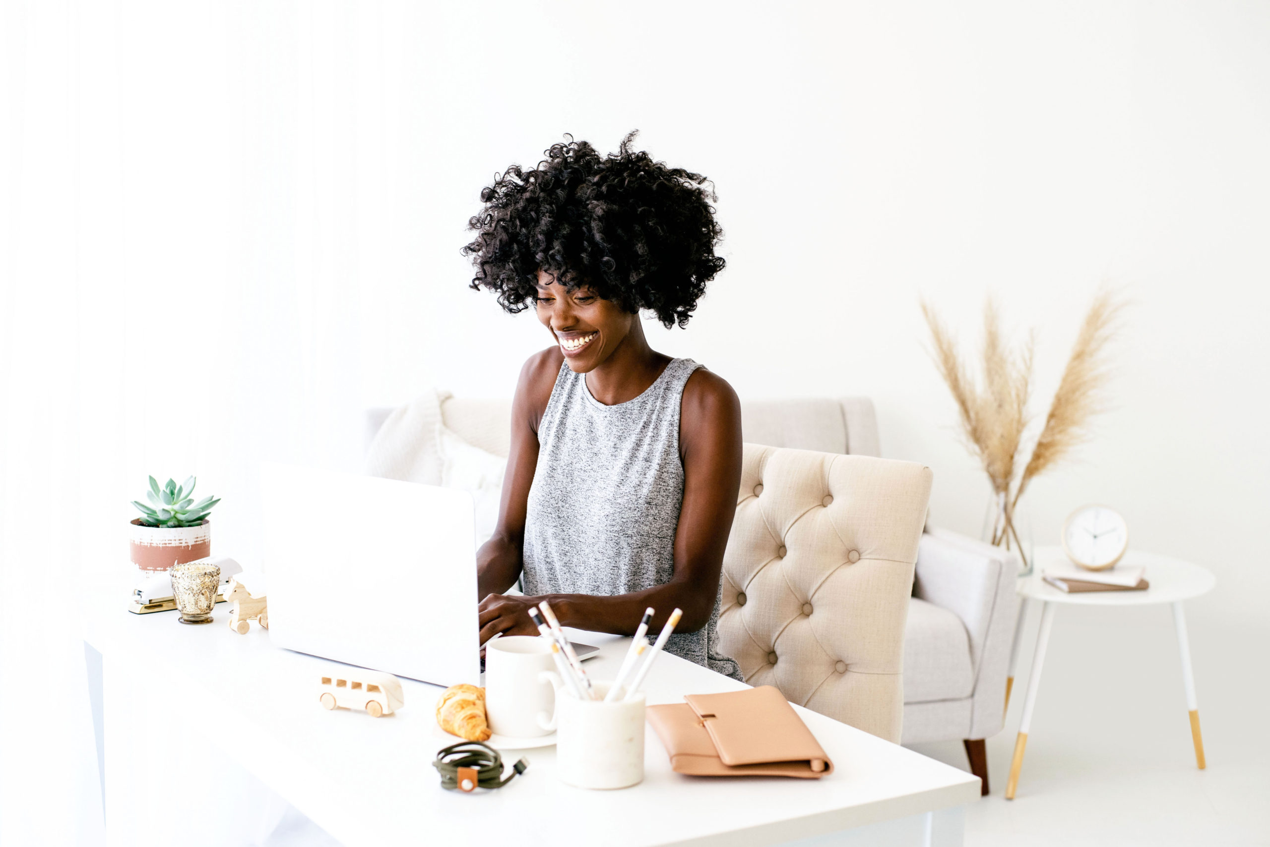 Woman sitting on a cream-colored chair, typing on her laptop that is sitting on a white desk.