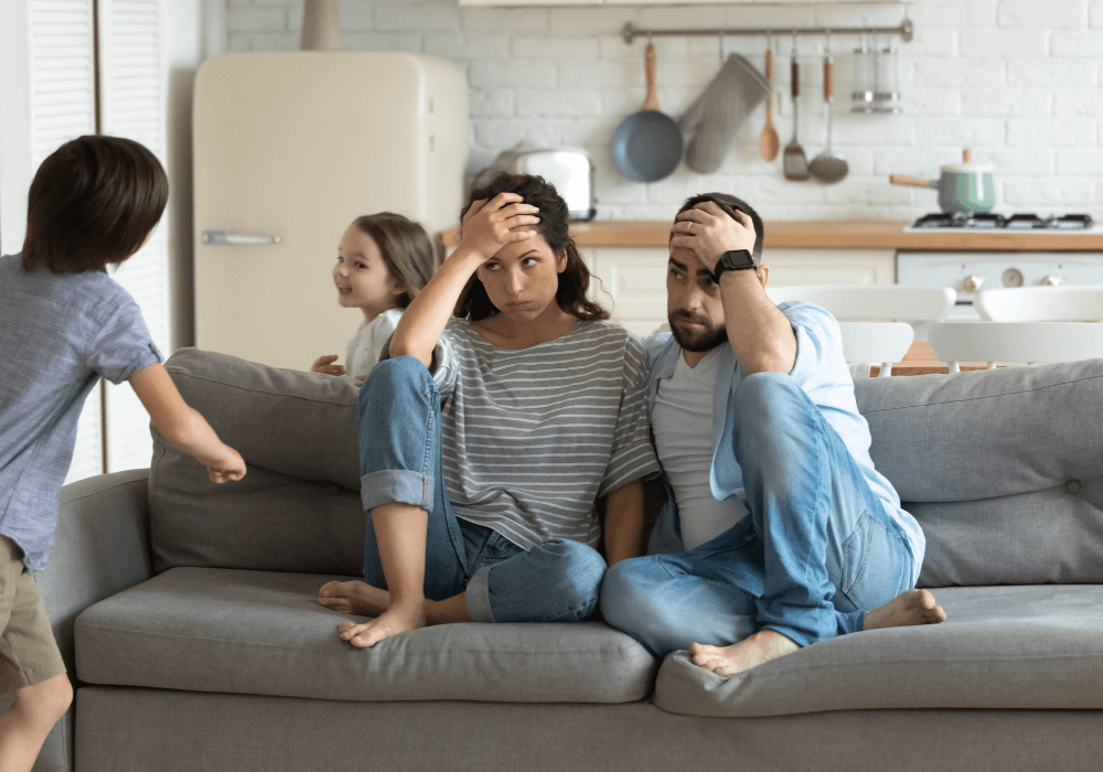 Two adults experience parental burnout while sitting on their couch watching their kids run around.